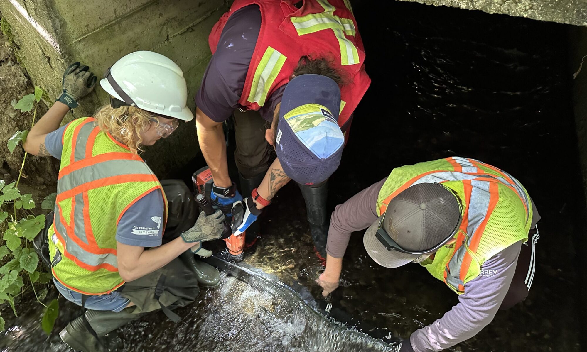 Team of workers in hard hats and high-visibility vests kneel at a dark, water-filled tunnel entrance, inspecting the area.