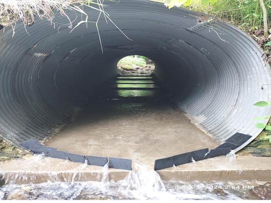 Large corrugated metal culvert with water flowing into the tunnel, light-visible opening at the far end and wet ground at the mouth.